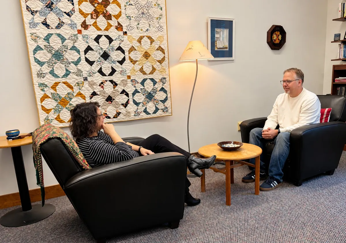 Rev. Ryan Irmer in a spiritual direction conversation, seated across a small table from a visitor in a quiet, lamp-lit room with a quilt on the wall.